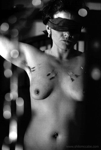 Blindfolded, female-bodied person with chest piercings, arms raised can be seen through some vertical hanging lengths of chain. 