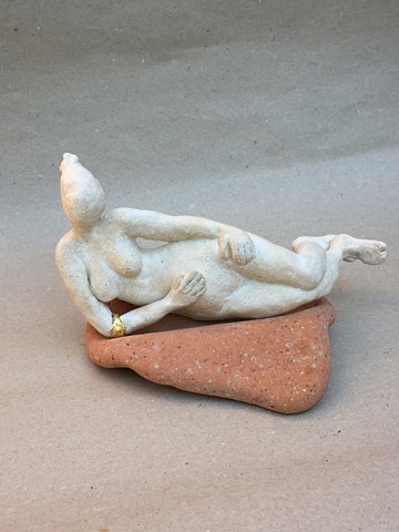 A small figurine, wearing a gold leaf bangle, sitting on a washed down ceramic tile, found on a beach 