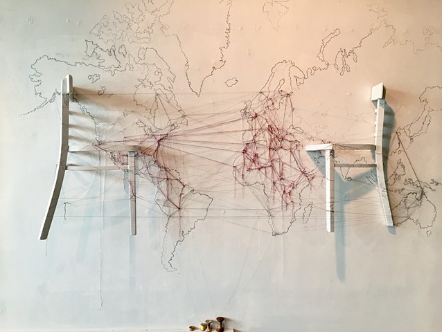 Thread from point to point on a map of the world traces major current movements of people.  A chair is split in half and secured to the wall on top of the graphite map of the world.