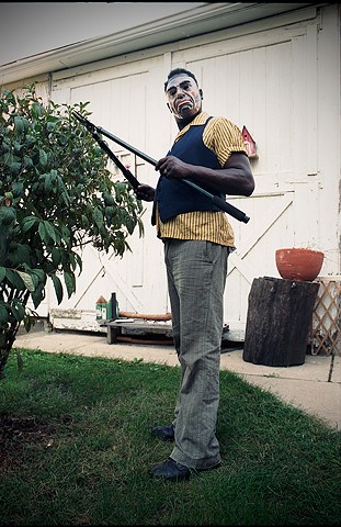 black middle aged man in scary mask that makes him look like a white man is pruning a shrub in his yard photographed by Lucy Mueller Photography