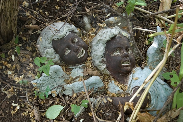 Plaster angels painted with black faces are buried in  overgrowth photographed by Lucy mueller