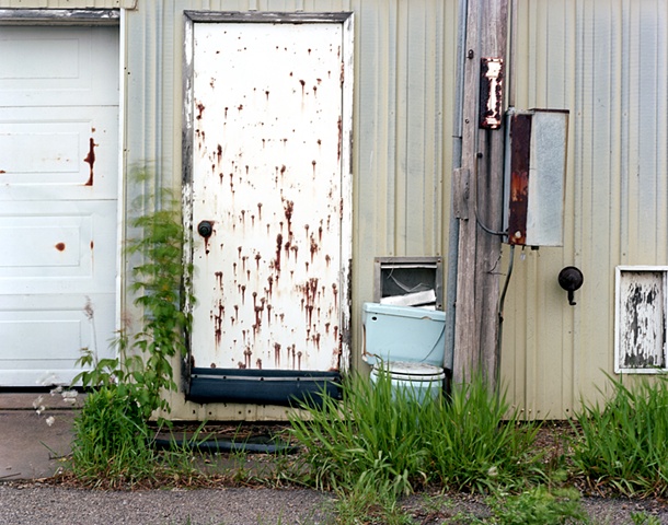 image of rust spotted barn with toilet by a door in grass.