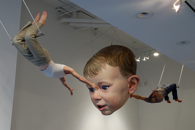 Figurative, polychrome sculpture of a life-size headless woman and man tossing a huge toddler's head between them on a trapeze