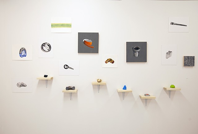 a number of small paintings installed on a wall above shelves at various heights holding small sculptures