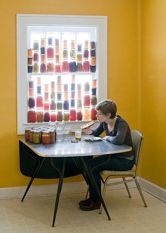 Self portrait in yellow room eating toast. Window is filled with canning jars full of jam, preserves. 