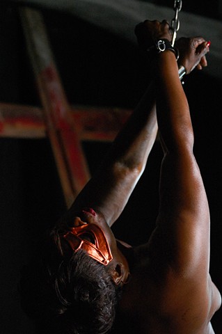 black woman with orange mask has her wrist shackled in the dungeon with a cross in the background