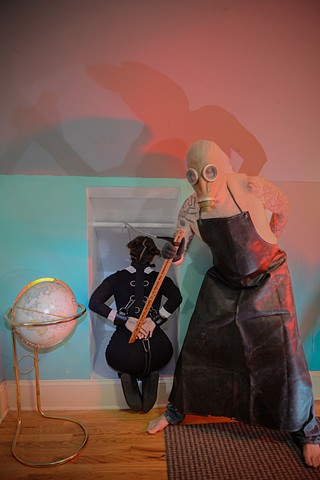 a man in an gas mask and butchers apron keeps a young girl captive in an alcove photographed by La Mouge Photography using colored lighting 