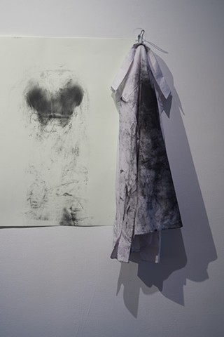 a white button up shirt hangs from a hook at the edge of a large drawing. The back of the shirt is dirtied black. The drawing is a pressed shape of lungs or maybe shoulder blades.