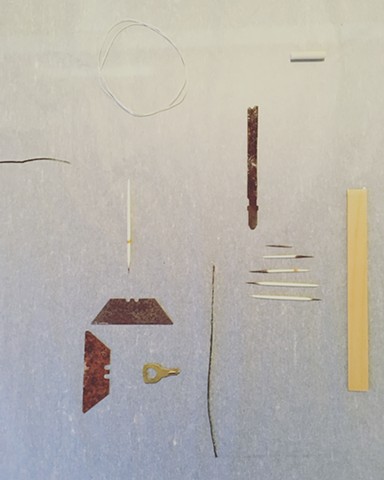 Objects on a paper surface. Two rusty razor blades are toward the bottom left corner; one is vertical with the sharp edge on the left, and the other is horizontal with the sharp edge pointing up. They create half an open square. Framed by this shape is a 