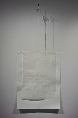 A nebulizer mask hangs from the ceiling, it's shadow cast against the wall. The end of the tube reaches over an unframed drawing. It has two layers of paper, one much finer and smaller than the backdrop. The white surface is water marked, crumpled in some