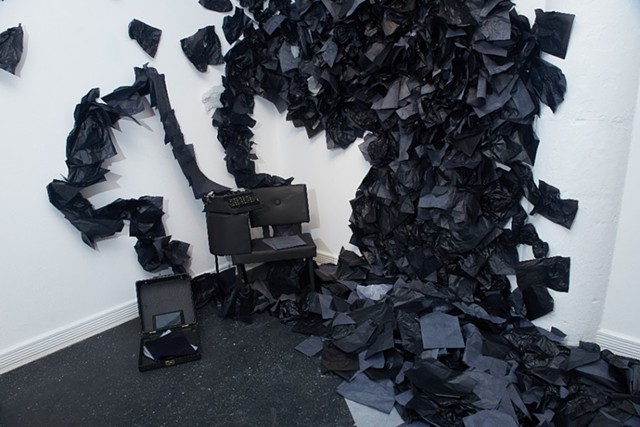A view from above of a large black and white installation. There is a chair with a small desk attached to it, painted in matte black. On the desk is an old typewriter. Coming from the typewriter is a sheet of black carbon paper that extends onto the wall 