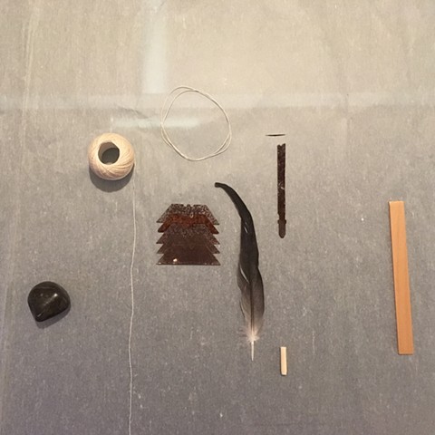 A cream colored piece of paper covers a surface. Arranged on this surface are found objects. To the left and towards the bottom corner of the image is a smooth black stone. Above that and to the right is a small ball of white string that is a little unwou