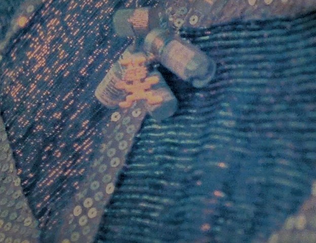 Close-up of golden sequin and black stripped pattern fabric and a golden Precolombian figurine and several small glass bottles at the top of the image. The frame is a film still and the golden objects all appear to have a bluish tint.