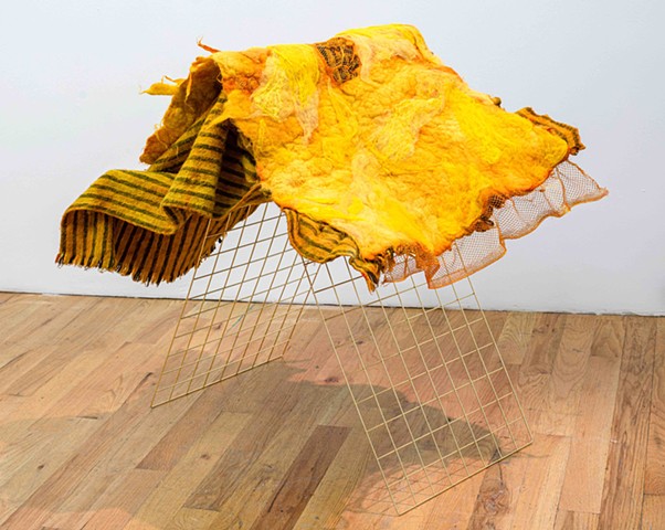 A two-layer textile sits atop a metal grid structure on a wood floor against a white wall. the grid is gold and made of two flat panels leaning up against one another. The bottom layer of the textile is a yellow panel with grey stripes and three black str