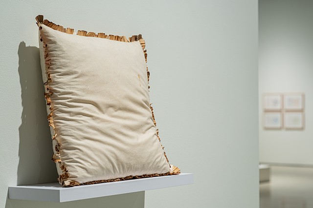 A square throw pillow sits upright on a white shelf against a gallery wall seen from a 3/4ths view. The pillow is off-white with marks from use. Wrapped around the entirety of the seams are unraveled and pleated joint filters sewn into the pillow. They ar