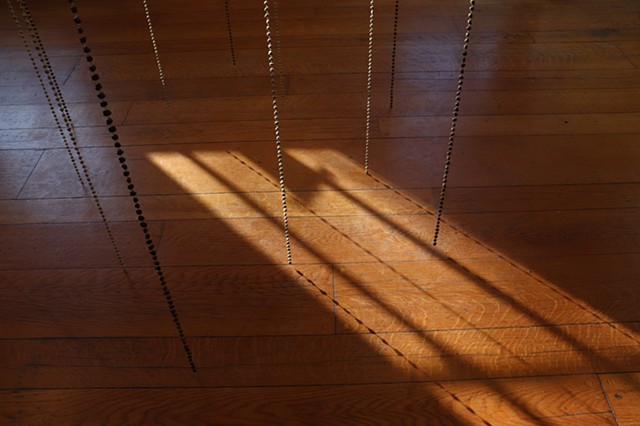 A detail image pointed at the floor shows more clearly that there is an inner ring of unroasted coffee beans and an outer ring of roasted beans and space to stand in the middle of the installation of brass chains of coffee beans hung from the ceiling. Warm sunlight tenderly casts their shadows on the wood floor.