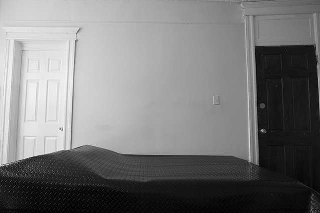 A grey-scale photograph. Black diamond plated rubber flooring lies on top of a bed, The bed is horizontal in the frame, and no bed frame can be seen. On the left of the image, the covered bed.bulges where pillows are usually placed. It is against a bedroo