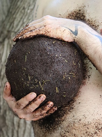 A vertical photograph of a close up of the artist's light skinned hairy belly with a sphere of moist dark coffee grounds with bits of green tea leaves sticking out, held between their hands. Their hand and arm on top of the ball of compost has a tattoo that looks like a squiggle of smoke. The ball is roughly the size of a mini watermelon or a soccer ball, and their hands, chest, belly and hip are covered in the grounds. The artist stands at an angle, as if holding part of their belly or something emerging from their belly. In the background we see textured tree bark.