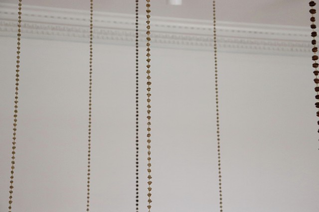 A detail shot of several coffee beans chains hung from the ceiling in front of a white wall, while symmetrically installed, here they appear staggered with some closer or farther apart. The focus is on a central unroasted coffee bean chain which has a pale green jade-like color. The coffee beans have been paired, flat sides together, so that they become beads. Reminiscent of a rosary or prayer beads, each bean has been drilled through and the pairs connected by small loops of golden brass wire.