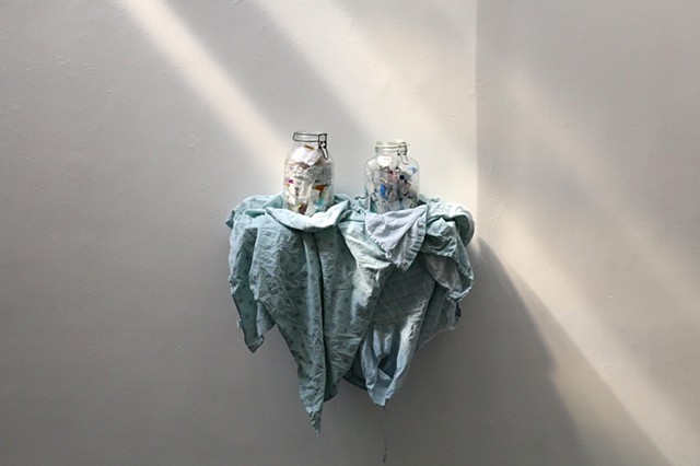Two glass jars sit side by side on a shelf draped with medical gowns. The jars contain an array of medical ephemera including used needles and prescription bottles. The piece is installed on a white wall. Coming in like a dream from the upper left at an a