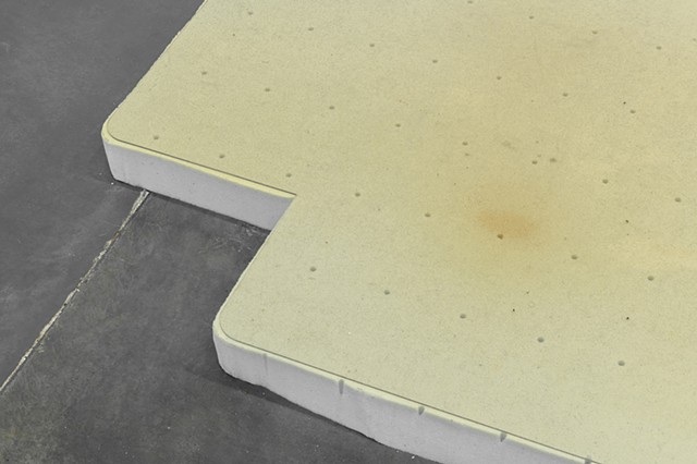 A memory foam mattress topper lies flat on a dark gray floor. On top of the memory foam is a thin clear plastic office chair mat with a lip. The image shows a close up cropped view of the lip which is a rectangular shape protruding from one edge of the ma