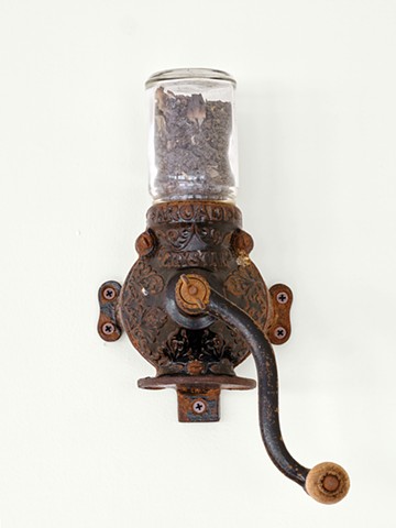 An antique coffee grinder is mounted on a white wall. It’s dark brown, covered in rust and has a rounded body with flanges protruding from the sides with matching rust color screws. A curved crank with a wood handle extends from the center of the coffee g