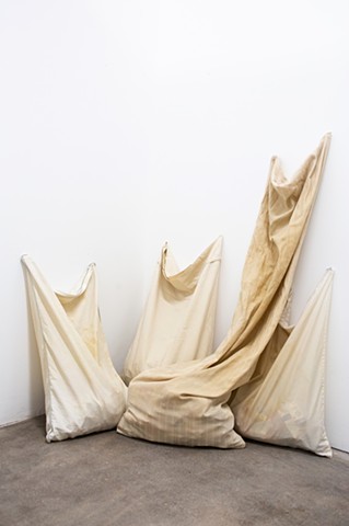 Four pillowcases, stained from use, sit vertically huddled together in a corner, on a brownish grey concrete floor. They lean against white walls, reaching upwards. One pillowcase is the size of a long body pillow. They are filled with medical ephemera wh