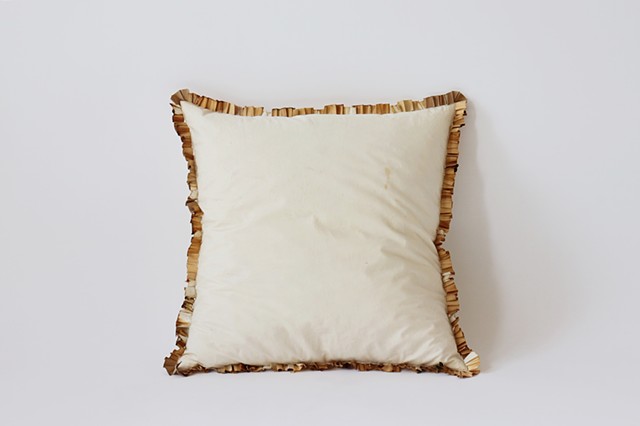 A square throw pillow lies upright against a white background. The pillow is off white with marks from use. Wrapped around the entirety of the seams are unraveled and pleated joint filters sewn into the pillow. They are a range of sh