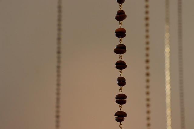 A close up shot of a chain of roasted coffee beans with more chains in the background. The coffee beans have been paired, flat sides together, so that they become beads. Reminiscent of a rosary or prayer beads, each bean has been drilled through and the pairs connected by small loops of golden brass wire.