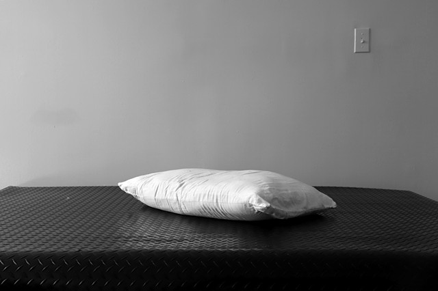 A grey-scale photograph. Black diamond plate rubber flooring lies on top of a bed. The crop of the frame cuts off the bottom and ends of the bed turning it into an ambiguous shiny textured surface. On top lies a pillow and the background only contains a 