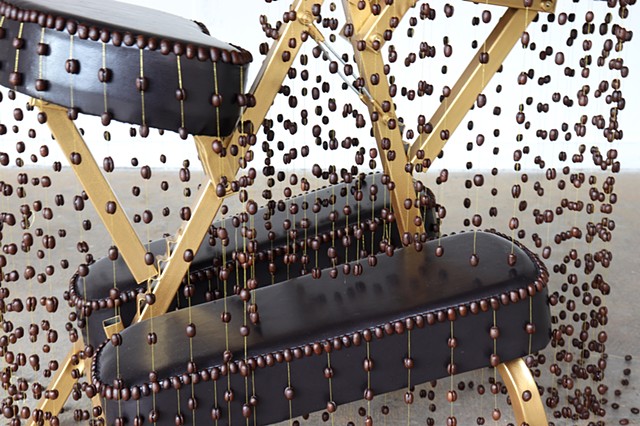 A close up of the middle of the chair's adjustable structure shows the coffee bean strands hovering above the upholstered legs of the chair and also shows how the beans are glued together in pairs on the gold threads.
