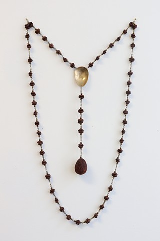 An abstracted rosary hangs from a white wall. The structure and counting system remains the same however pairs of coffee beans, drilled into and paired flat side together, make up each bead. Small round bronze color beads are placed on either end of the coffee bean pairs which are connected to each other and short lengths of chain by metal small loops. In place of a crucifix is an egg-shaped avocado seed. In place of the center piece that often depicts the Virgin Mary is an imageless head of a golden antique spoon. The work is hung by points along the prayer beads so that the spoon head and avocado pit hang in the center while the rest of the beads hang below in a long inverted arch.
