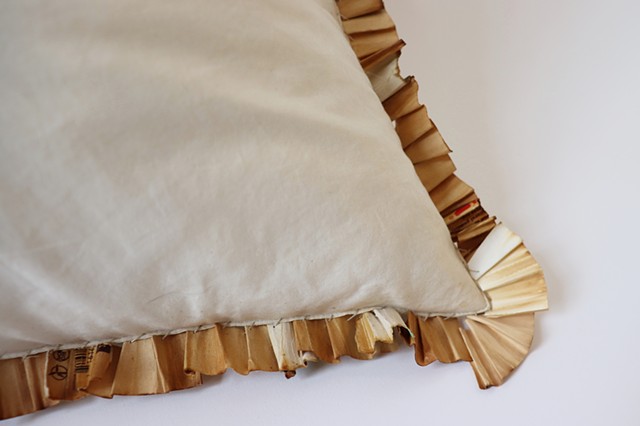 A close up of the bottom right corner of an off-white square throw pillow without a pillow case lies against a white background. Sewn into the seams of the pillow are unraveled and pleated joint filters. They are a range of shades, white, tan and differen