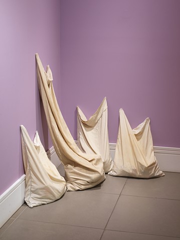 Four pillowcases, stained from use, sit vertically huddled together in a corner, on a grey tile floor. They reach upwards and lean against purple walls with a white trim at the bottom. One pillowcase is the size of a long body pillow. They are filled with