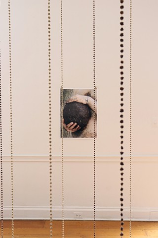 A photograph on a white wall of my bare torso holding a soccer ball sized ball of coffee grounds that looks like soil between my hands. My stomach is coated in the grounds and the placement of my hands mimic holding one's own pregnant belly. In the foreground several brass chains of green unroasted and brown roasted coffee beans hang down from the ceiling.