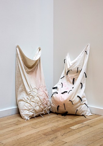 Two pillowcases sit in a corner on a wood floor against a white wall. Both are white with pale red stains, the left has injection needles (with caps) stuck through the bottom half of the fabric and the right has short strands of black yarn speckling the 