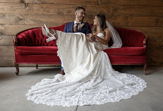 A bride and groom smiling at each other while sitting on a red couch with wood panel walls behind