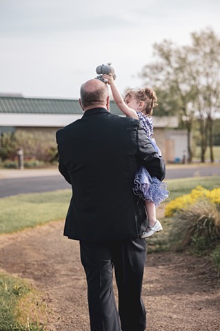 A grandfather walking away from the camera carrying his granddaughter who is placing her stuffed elephant atop his head
