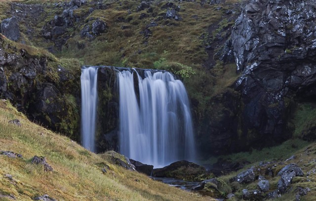 A waterfall with smooth flowing water in Iceland with green grass and black rock around