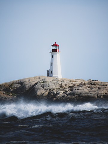 The lighthouse at Peggy's Cove against a blue sky with mountainous blue waves with white caps crashing in the foreground