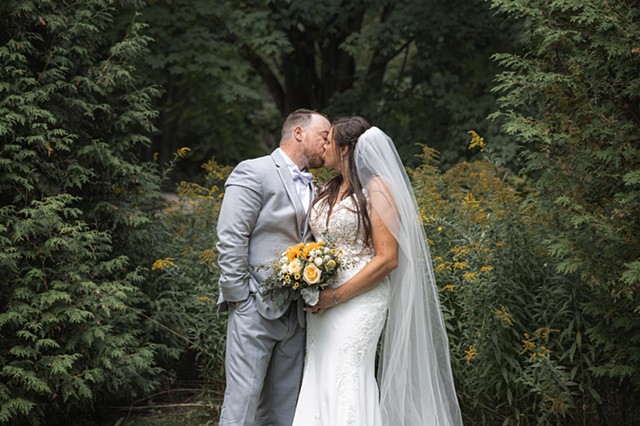 A bride and groom kissing between evergreens with yellow flowers and more trees in the background
