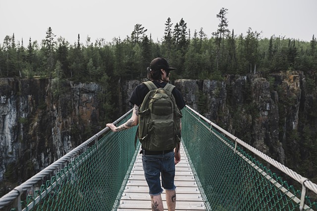 A man standing on a suspension bridge with a cliff face covered in moss with evergreen trees on top in Thunder Bay, Ontario
