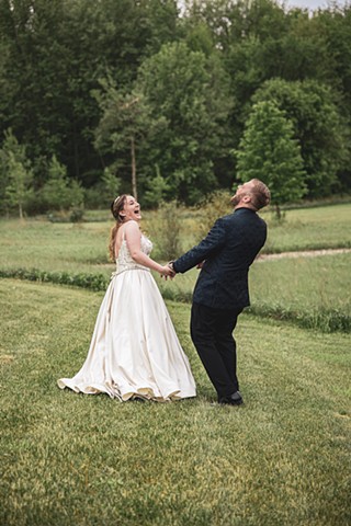 Bride and groom holding hand in field surrounded by trees laughing hard