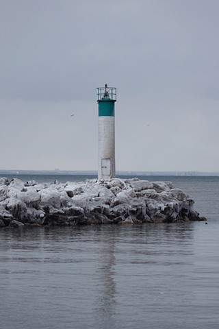 A white and blue lighthouse next to Lake Ontario covered in snow and ice with sea gulls flying around it