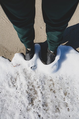 Legs wearing Roots Canada sweatpants, wearing Blundstone boots, with ocean waves on a sandy beach in Salisbury, Massachusetts