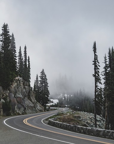 A winding highway in Mount Rainier National Park with cloud cover misting over evergreen trees in the background
