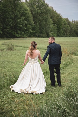 Bride and groom walking hand in hand in a field lined with big green trees