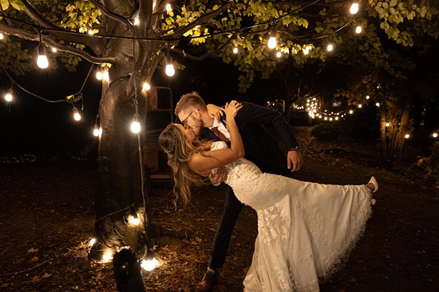 A groom dipping his bride and kissing her in front of a tree and path at night illuminated by twinkling lights