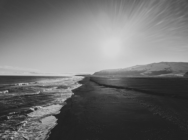 A black and white photo of the ocean and black sand on the south coast of Iceland with mountains to the side