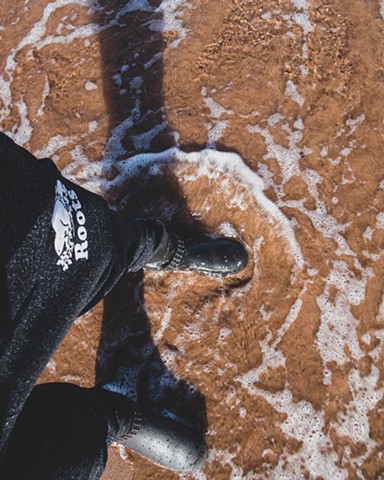 A person wearing Roots sweatpants and Hunter boots with a red sand beach and ocean water below